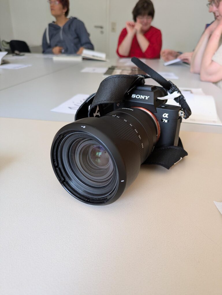 Das Bild zeigt eine Kamera, die auf dem Tisch liegt., Im Hintergrund sind Menschen zu erkennen, die auch an dem Tisch sitzen. Der Zweck des Bildes ist: ein Kurs hat stattgefunden. Der Kurs hat Fotos zum Thema gehabt. 