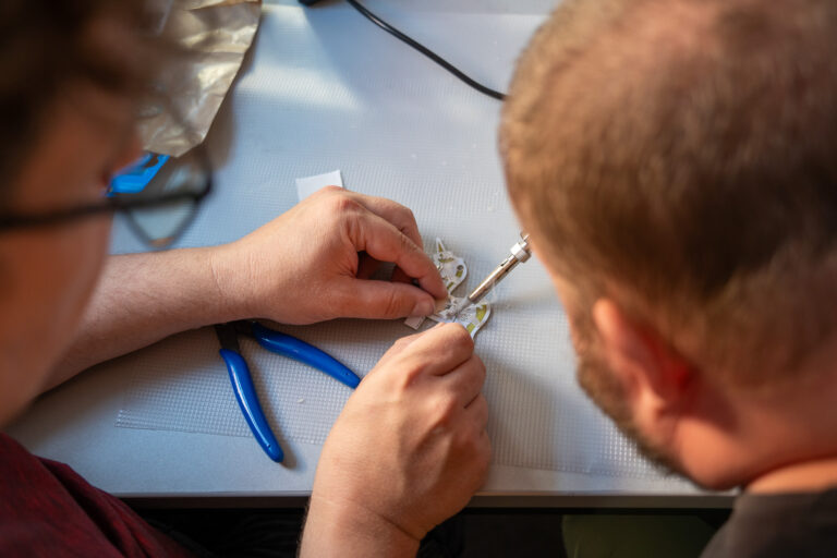 Das Bild zeigt, wie etwas repariert wird. Es liegt eine Zange auf dem Tisch. Die beiden Menschen, die von hinten zu sehen sind, löten gerade etwas zusammen.