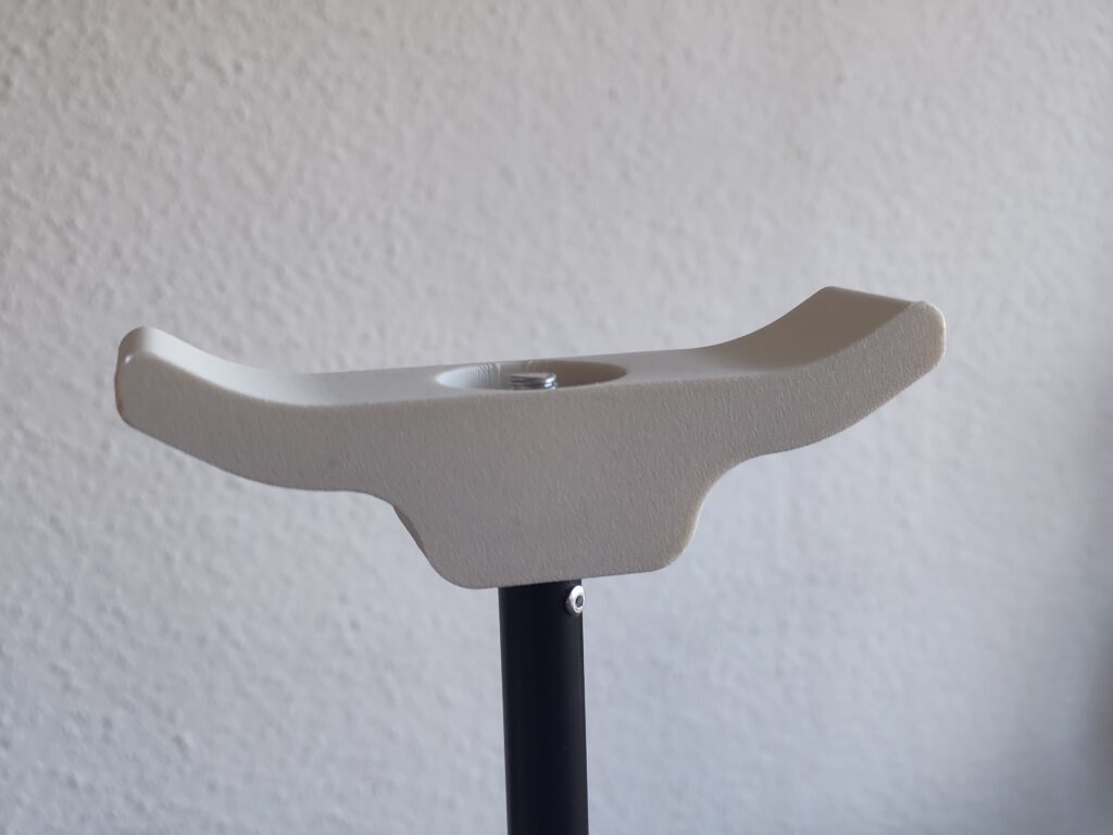 Das Bild zeigt eine Kinnauflage. Sie ist auf einem Metallstab aufgeschraubt. Die Kinnauflage ist beige. der Metallstab schwarz. Die Konstruktion dient dazu, das Kinn darauf aufzulegen, um eine Nahaufnahme des Auges, speziell der Regenbogenhaut aufzunehmen.